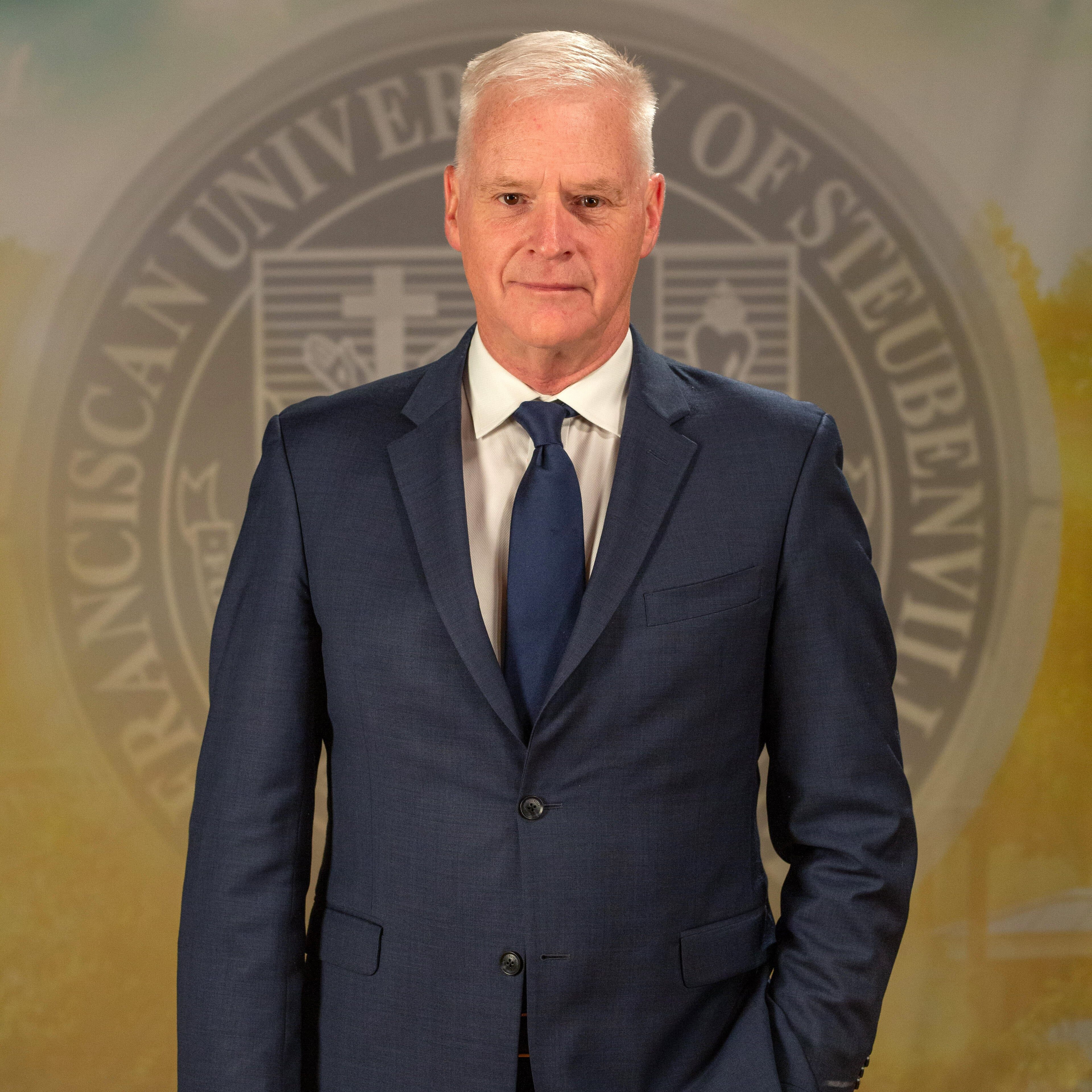 Stephen Lewarne_FNL__B5A2319 Dr. Stephan Lewarne in a navy suit standing in front of an university seal.