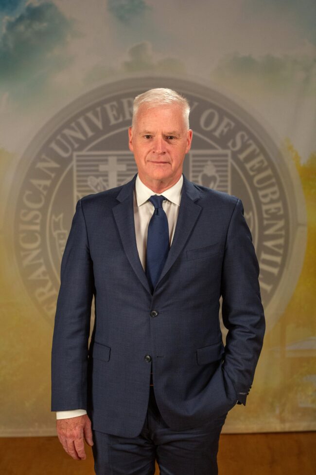 Stephen Lewarne_FNL__B5A2319 Dr. Stephan Lewarne in a navy suit standing in front of an university seal.