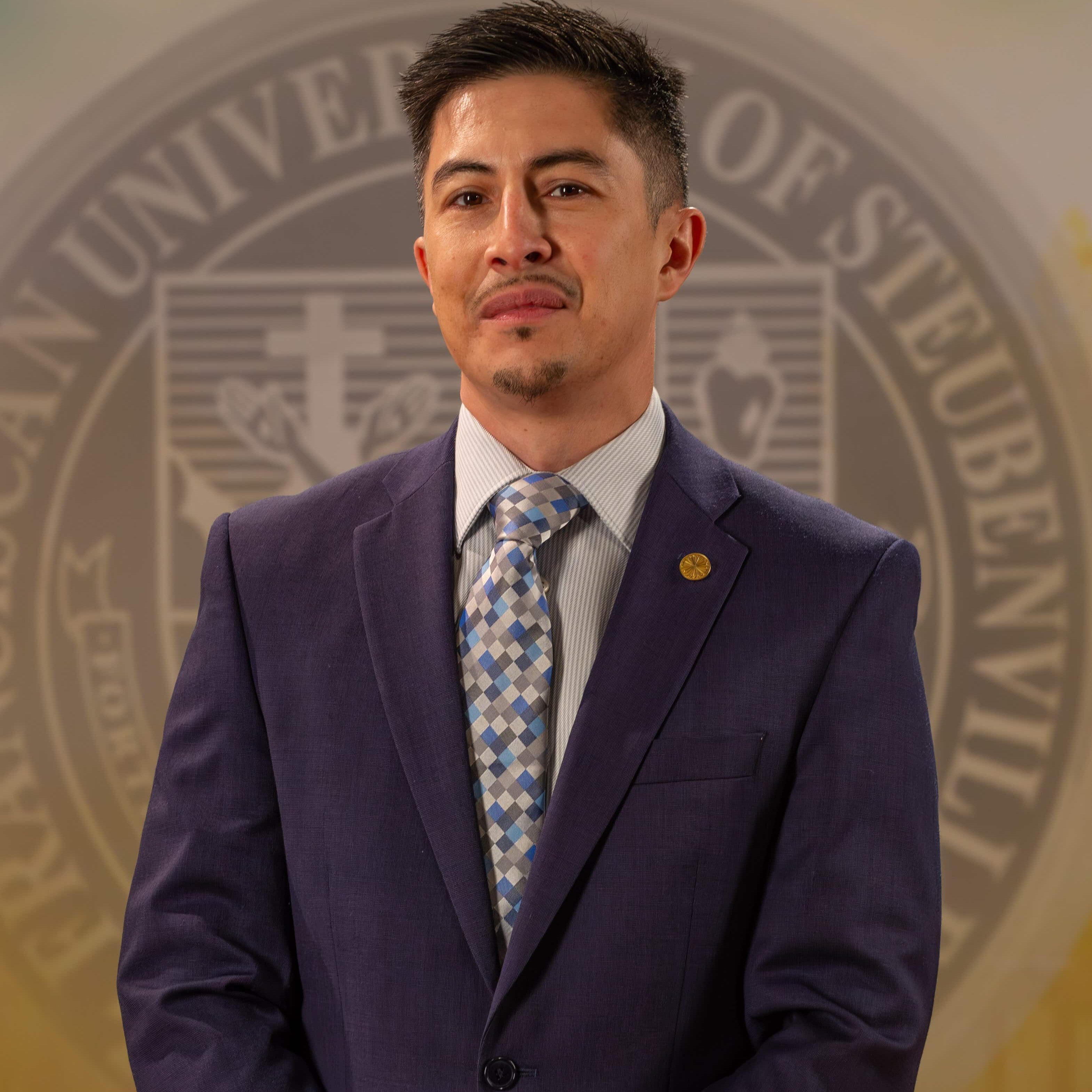 Dr. Matthew Escano_web portrait_FALL2025_ Dr. Matthew Escaño in a suit and tie standing in front of a university seal.