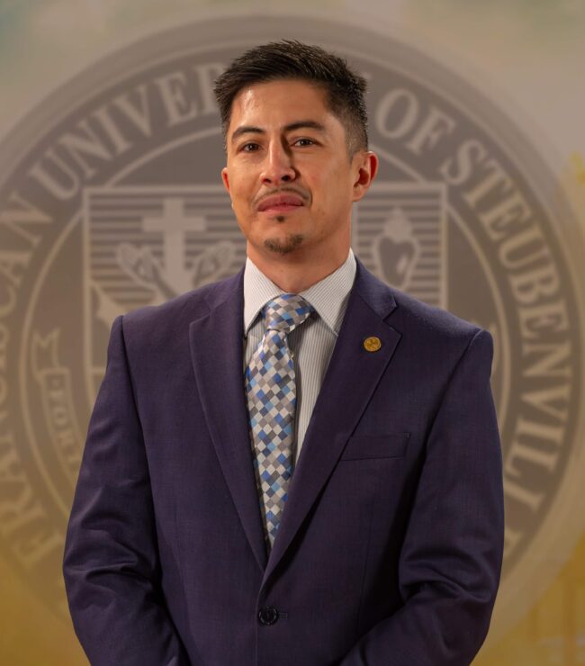 Dr. Matthew Escano_web portrait_FALL2025_ Dr. Matthew Escaño in a suit and tie standing in front of a university seal.